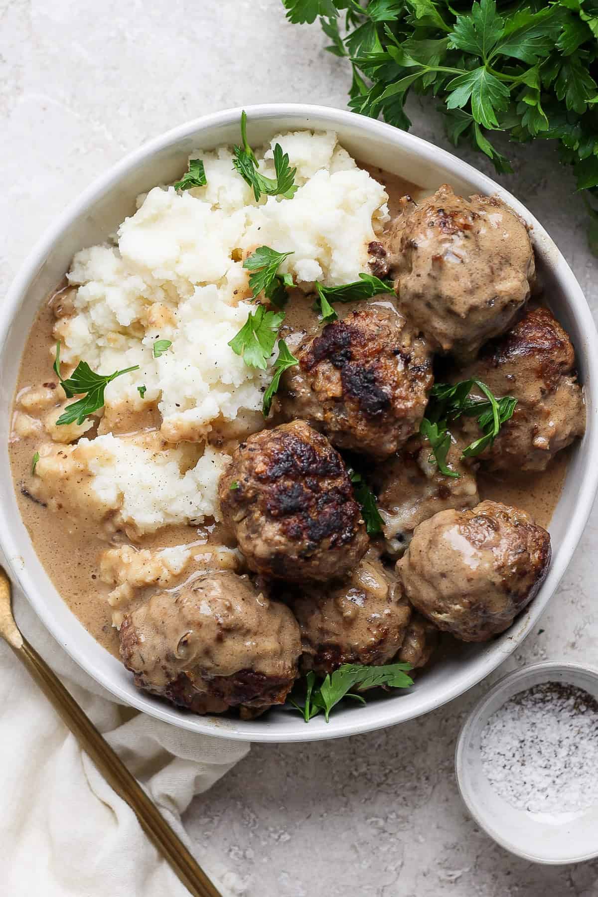 A plate of meatballs covered in gravy, served with mashed potatoes and garnished with fresh parsley. A fork and a small bowl of pepper sit adjacent to the plate.