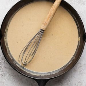 A cast iron skillet filled with a light brown sauce, with a whisk resting inside.