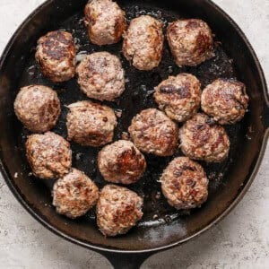 A cast-iron skillet contains 16 evenly browned meatballs on a textured light-colored surface.