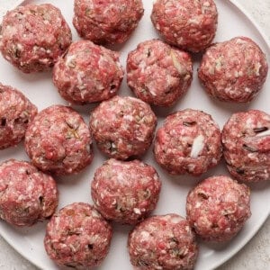 A plate filled with raw, evenly-sized meatballs, arranged in a circular pattern on a white surface.