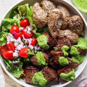 A bowl containing roasted potatoes, a fresh salad with lettuce, tomatoes, and onions, and air fryer steak bites topped with green sauce.