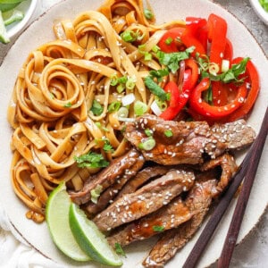 A plate with cooked noodles, sliced red bell peppers, and slices of beef topped with sesame seeds. A lime wedge is on the side, and a pair of chopsticks rests on the plate.