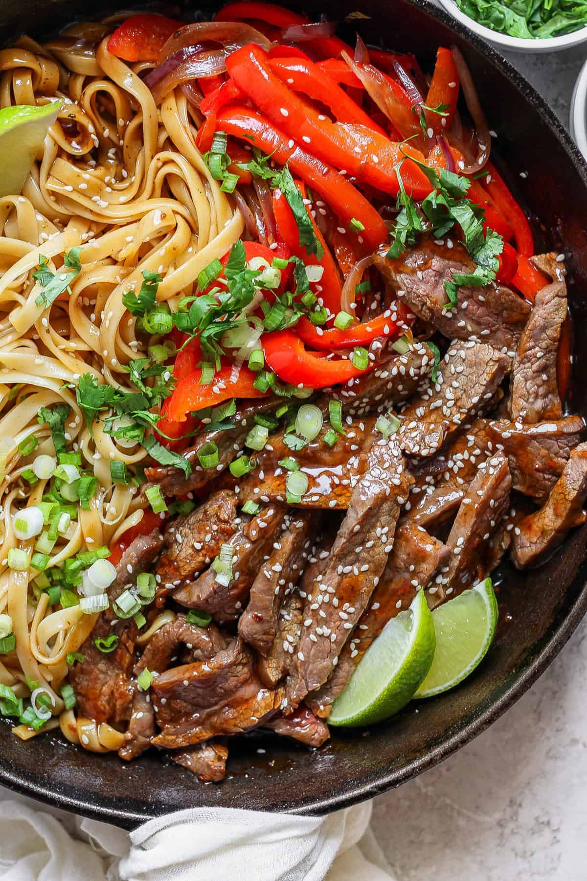 A bowl of stir-fried beef strips with sesame seeds, bell peppers, and noodles, garnished with chopped green onions and cilantro, alongside lime wedges.