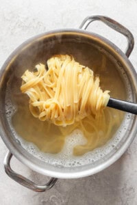 A pot of hot water with steam rising and cooked fettuccine noodles being lifted out using a spoon with slots.