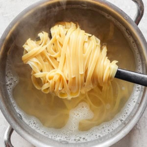 A pot of hot water with steam rising and cooked fettuccine noodles being lifted out using a spoon with slots.