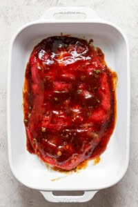 A raw marinated piece of meat lies in a white rectangular ceramic dish, coated with a reddish-brown sauce and garnished with herbs.