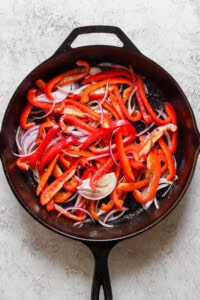 A cast iron skillet filled with sliced red bell peppers and red onions on a light-textured surface.