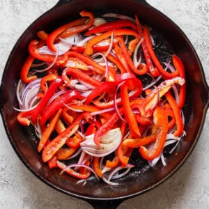 A cast iron skillet filled with sliced red bell peppers and red onions on a light-textured surface.