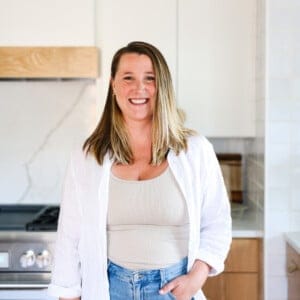 A woman with long hair, wearing a white shirt over a beige top and blue jeans, stands in a modern kitchen smiling at the camera, perhaps sharing her latest fit foodie finds.