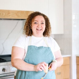 A curly-haired person, wearing a white t-shirt and a blue apron, is smiling while grating cheese in a kitchen, embodying the joy of culinary creation and foodie finds.