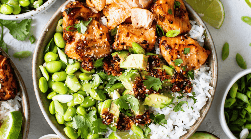 A bowl filled with grilled salmon, edamame, avocado, and white rice, garnished with sesame seeds and cilantro. Lime wedges are on the side.