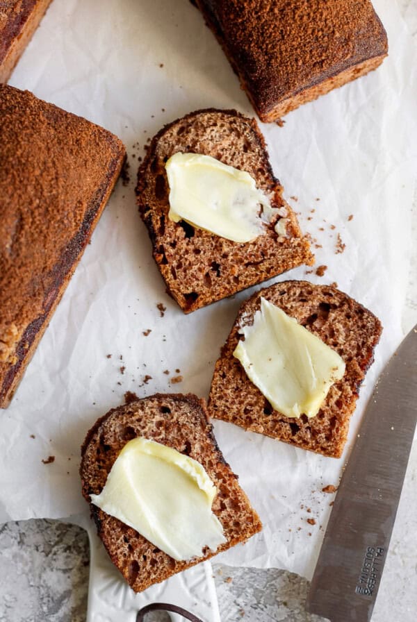 Two slices of dark bread with butter on a parchment paper, next to a knife with a partially visible bread loaf.