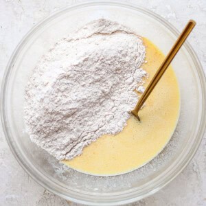 A clear bowl contains a mixture of liquid ingredients and dry flour with a gold spoon, set on a textured white surface.