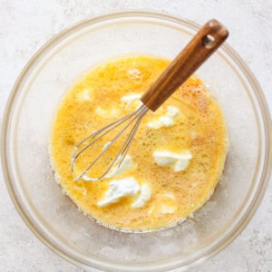 A glass bowl containing a beaten egg mixture and dollops of cream or butter, with a whisk resting inside.