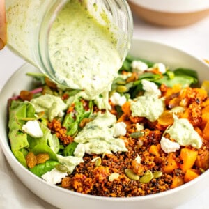 A hand pouring green herb dressing from a jar onto a bowl containing quinoa, roasted vegetables, greens, seeds, and crumbled cheese with another bowl of roasted vegetables in the background.