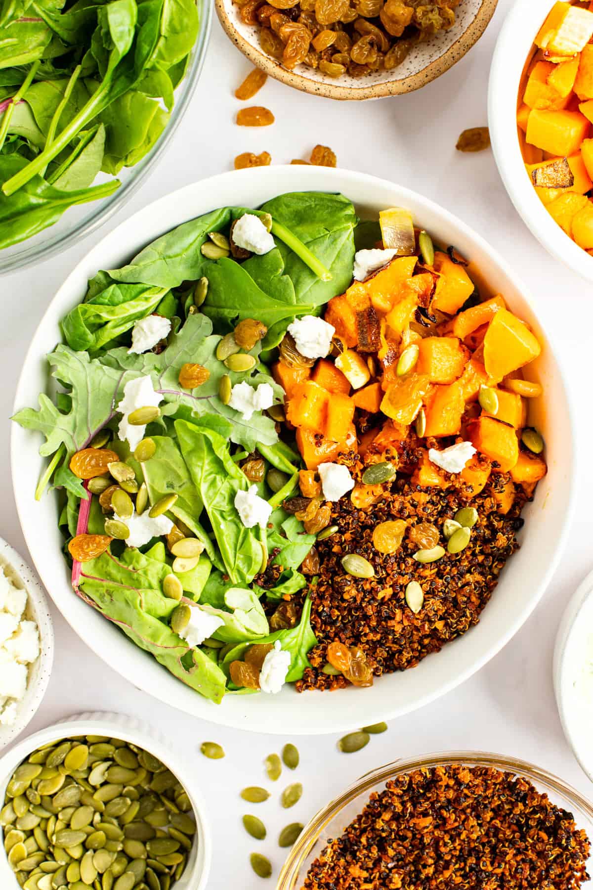 A bowl of salad with spinach, roasted butternut squash, quinoa, pumpkin seeds, and goat cheese crumbles, surrounded by small bowls of salad ingredients, including raisins and seeds.