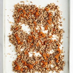 A baking tray covered with parchment paper holds a single layer of evenly spread, mixed quinoa with a toasted appearance.