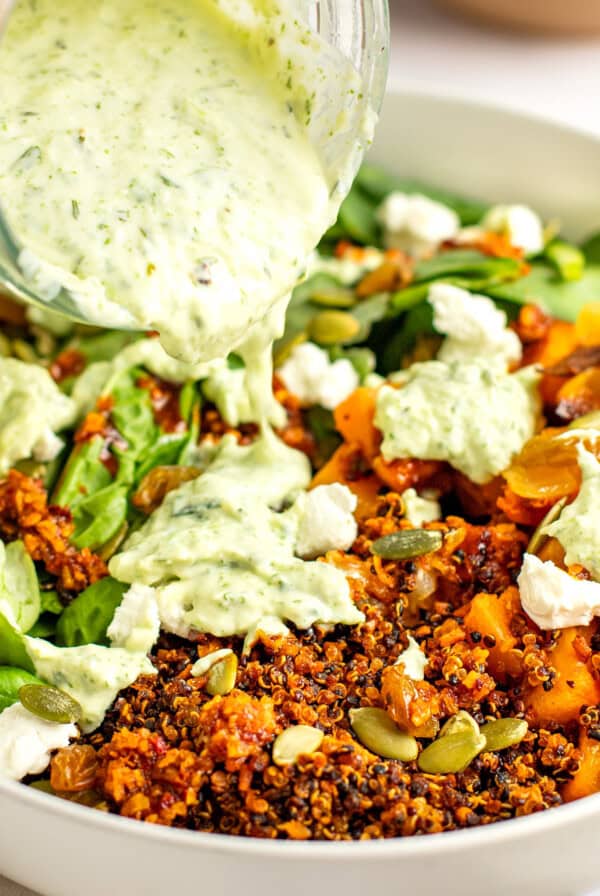 A close-up of a salad in a white bowl with quinoa, mixed greens, pumpkin seeds, raisins, and crumbled cheese, being topped with a creamy green dressing poured from a jar.