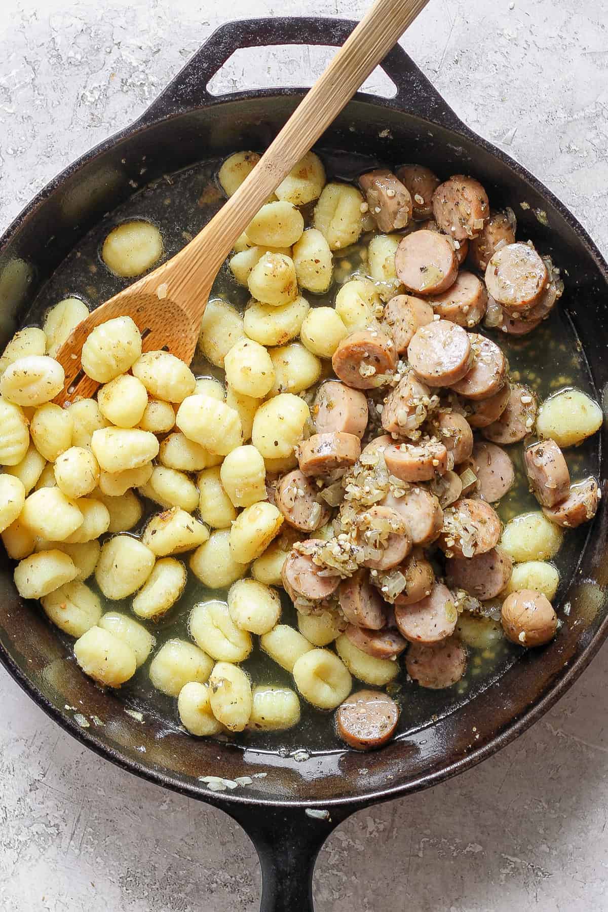 A skillet containing cooked gnocchi and sliced sausages mixed with a sauce, with a wooden spoon resting inside.