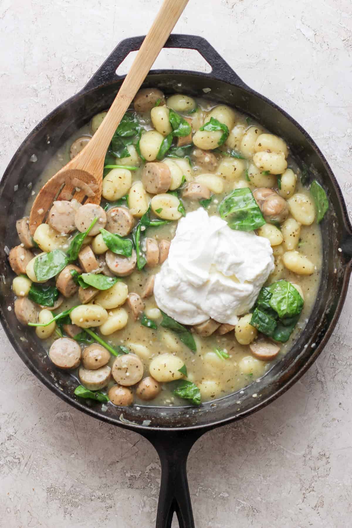 A skillet filled with gnocchi, spinach, sliced sausage, and a dollop of ricotta cheese, with a wooden spoon rested on the edge.