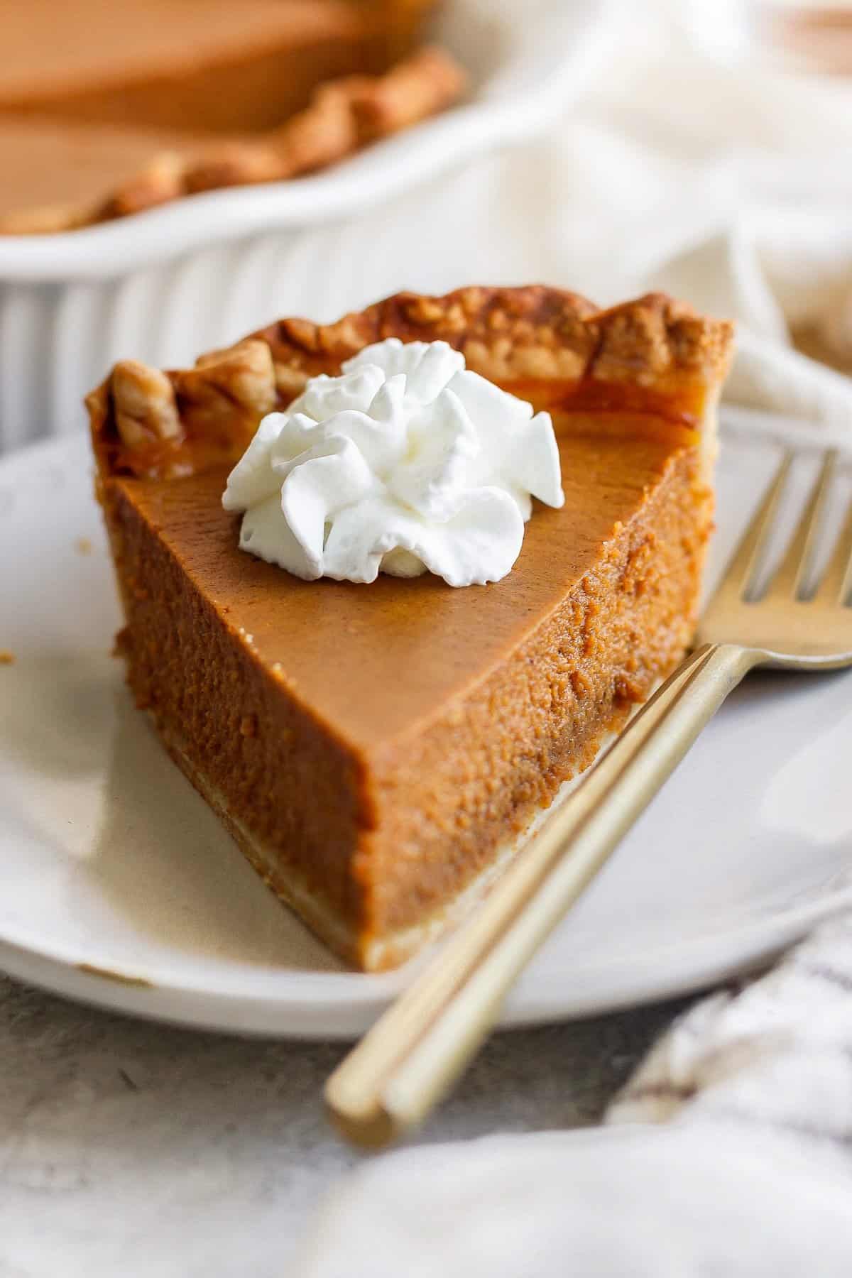 A slice of pumpkin pie with a dollop of whipped cream on top, placed on a white plate next to a fork.