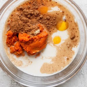 A glass bowl containing pumpkin puree, two eggs, milk, and various dry spices, ready to be mixed.