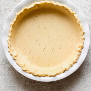 A pie crust in a white pie dish with a crimped edge and multiple small fork holes in the base, placed on a light textured surface.