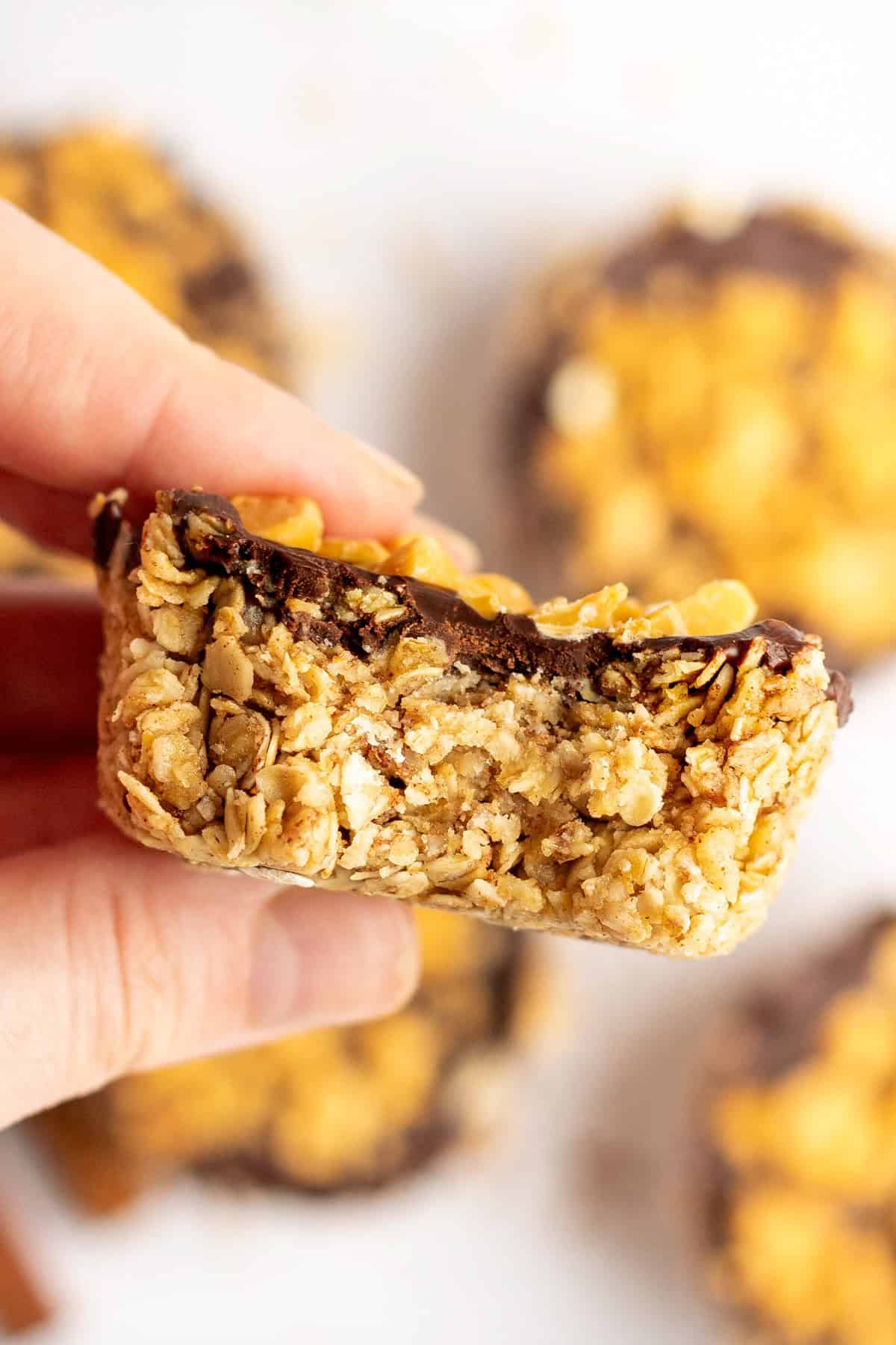 Close-up of a hand holding a homemade granola bar with a chocolate topping, several more out-of-focus granola bars are in the background.