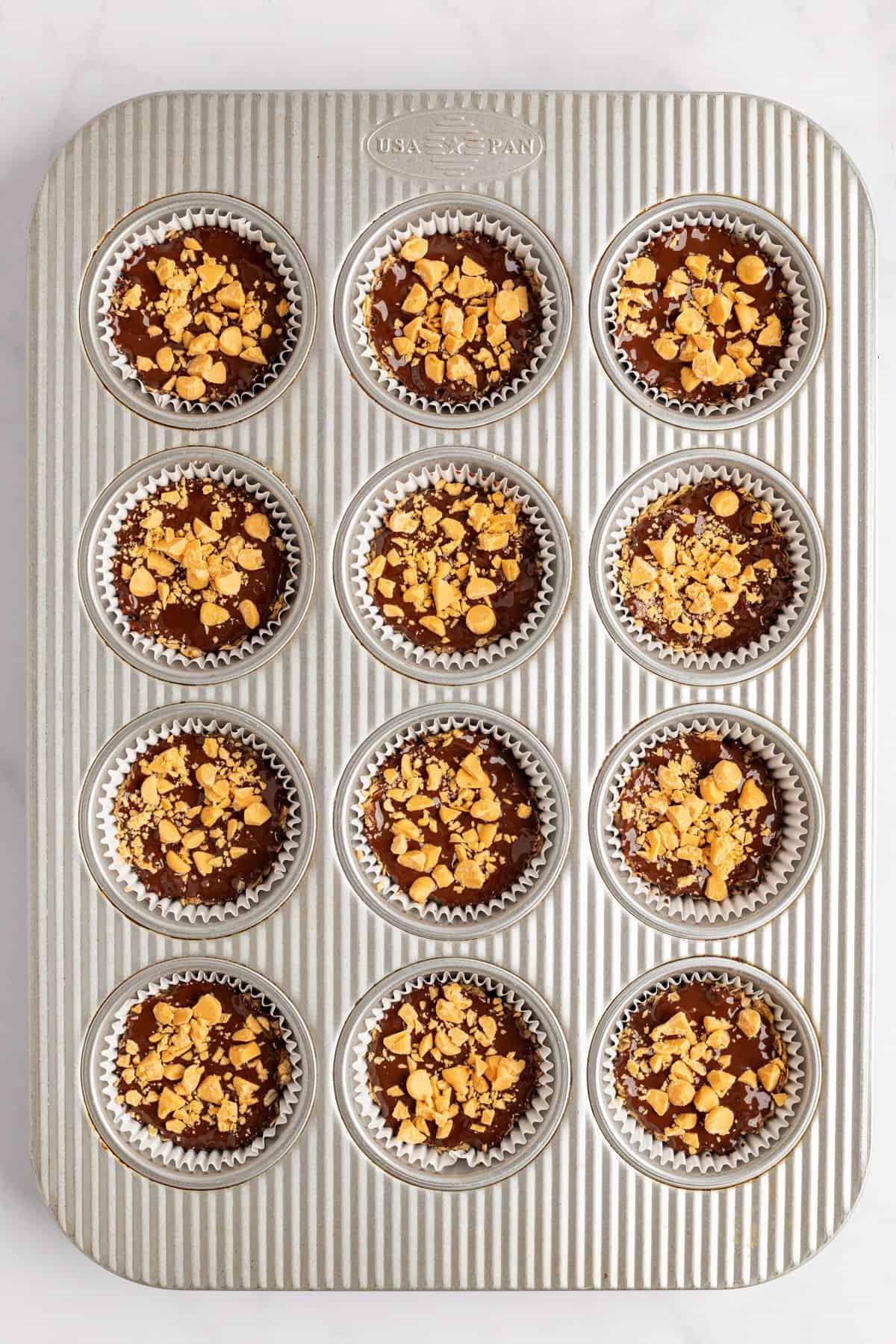 Muffin tin with twelve slots filled with chocolate batter topped with crushed nuts, lined with silver foil cupcake liners, ready for baking.