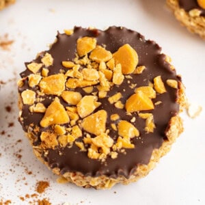 Close-up of a round dessert with a chocolate top layer sprinkled with chunks of yellow-orange toffee pieces, set against a light background.