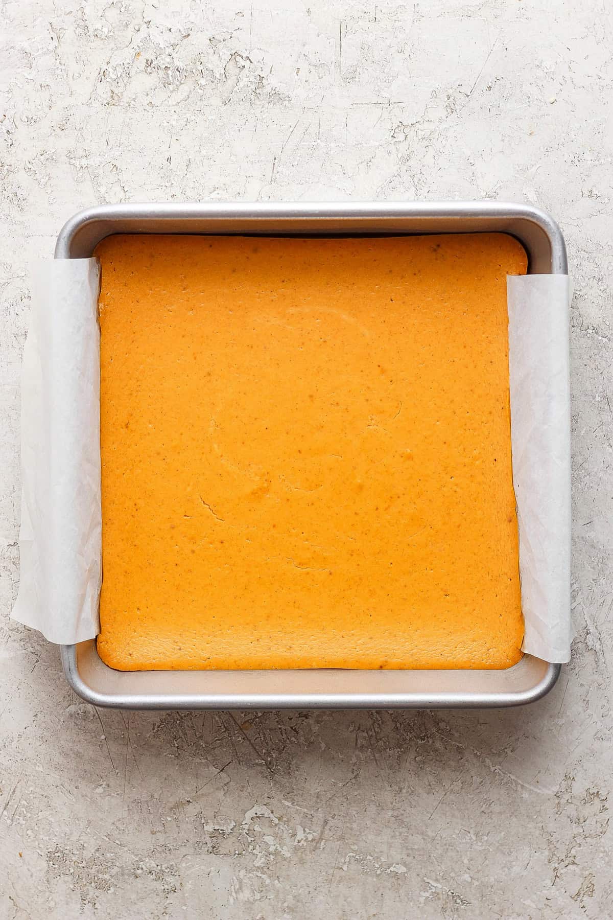 A square pan with a light golden-brown baked cake lined with parchment paper on a textured, light-colored surface.