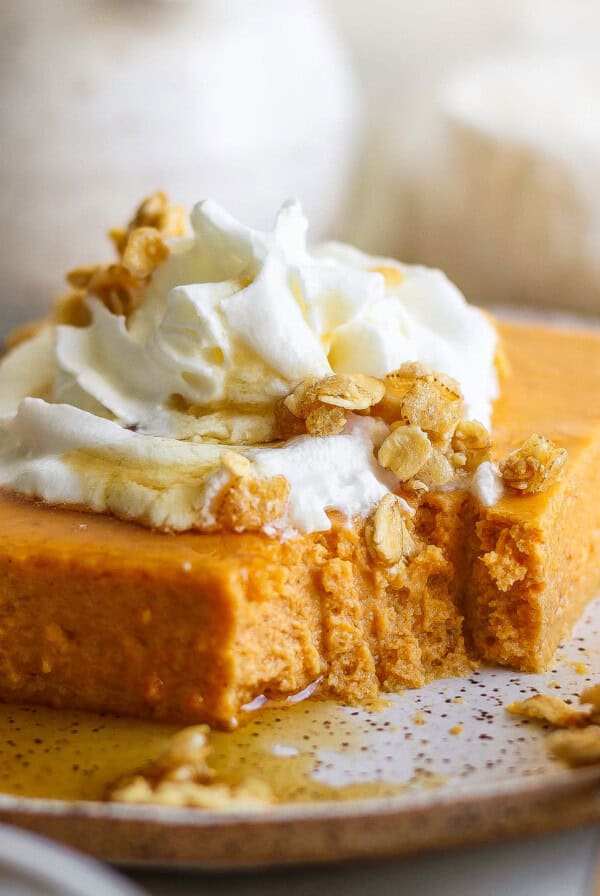 A slice of pumpkin dessert topped with whipped cream and granola on a plate, with a fork beside it. A small portion is missing from the dessert.