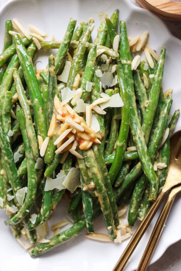 A plate of green beans topped with shaved Parmesan cheese and almond slivers, with serving utensils on the side.