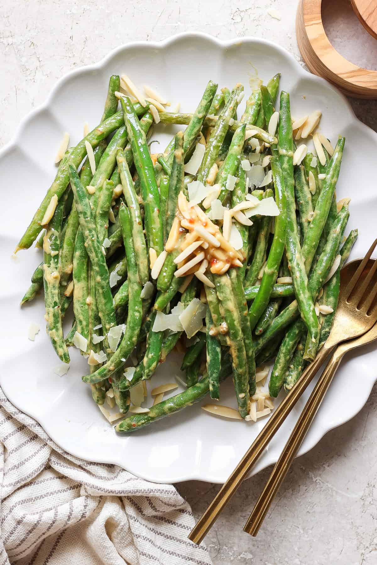 A plate of green beans topped with sliced almonds and shaved parmesan cheese, served with a gold fork and knife on a white tablecloth.