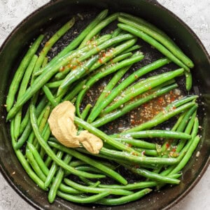 A cast iron skillet with cooked green beans, mustard, and red pepper flakes on a textured white surface.