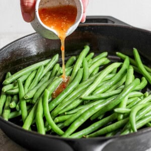 A hand pours a small container of orange sauce onto fresh green beans in a black cast iron skillet.