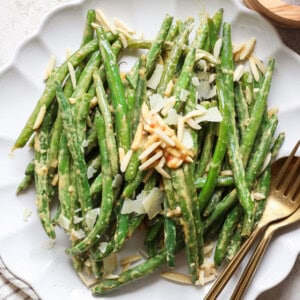 A plate of green beans topped with shaved almonds and cheese, accompanied by a fork and knife.