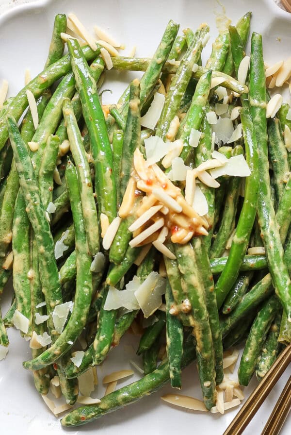 A plate of green beans topped with shaved almonds and cheese, accompanied by a fork and knife.