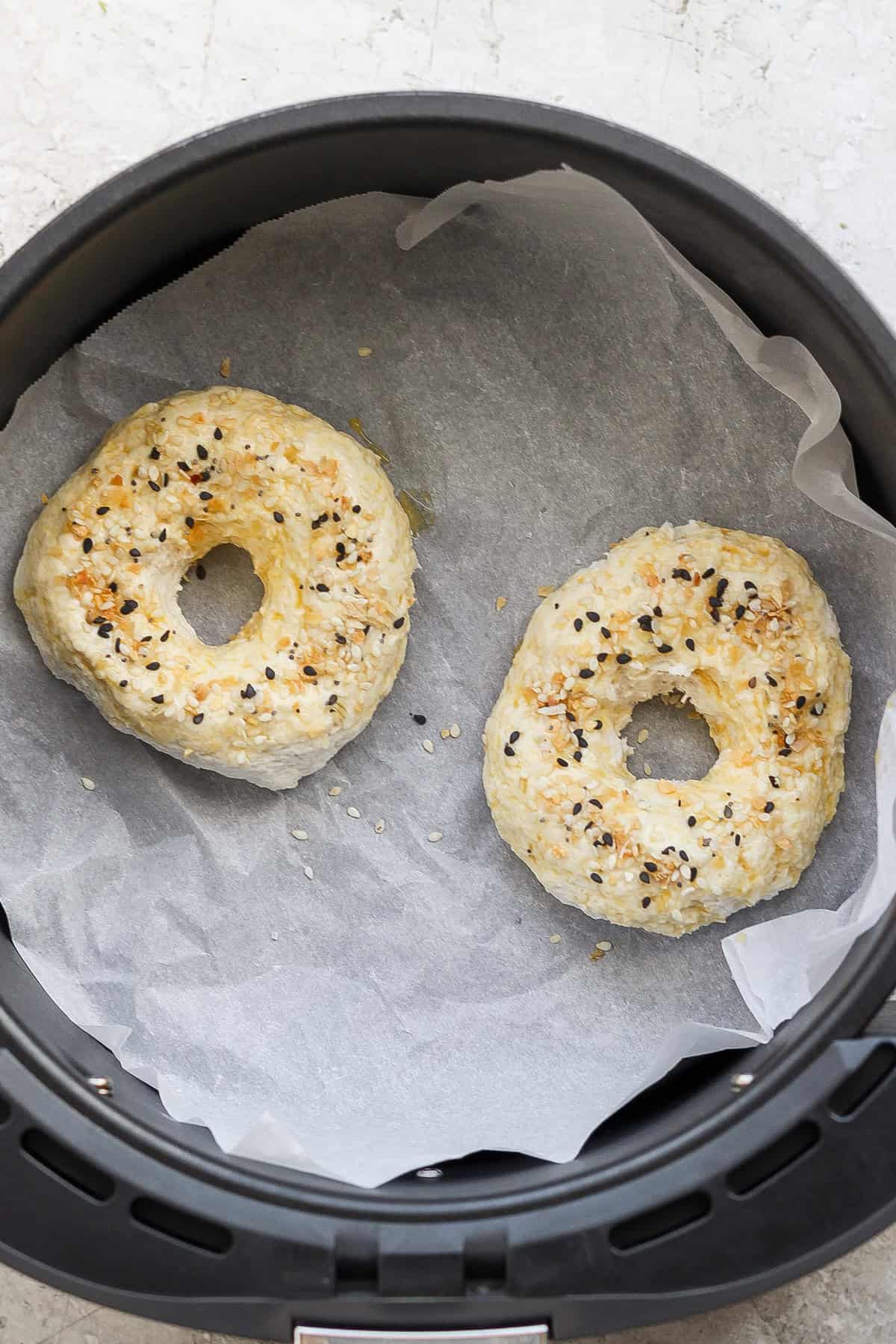 Two bagels with seasoning on top are being cooked on parchment paper inside an air fryer.