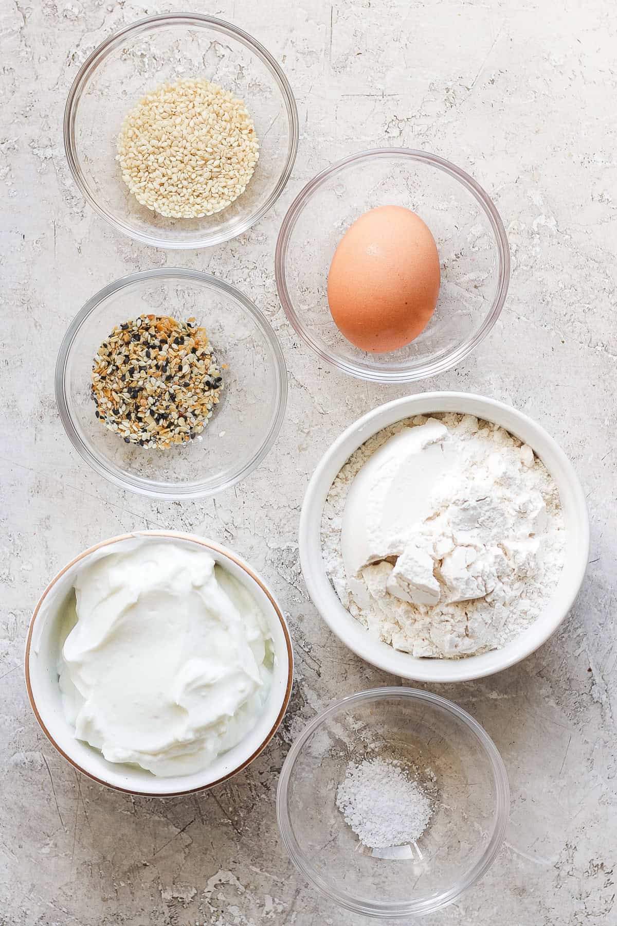 Six bowls containing various ingredients and one egg on a textured surface. The bowls hold sesame seeds, a spice blend, flour, a creamy substance, and salt.