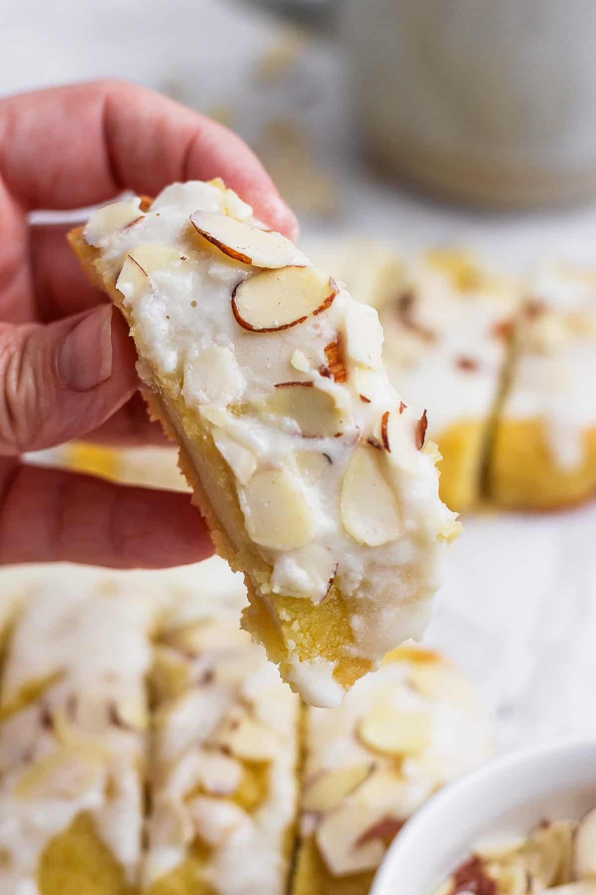 A hand holding a slice of almond-topped dessert bar with white icing. Other similar dessert bars are visible in the background.