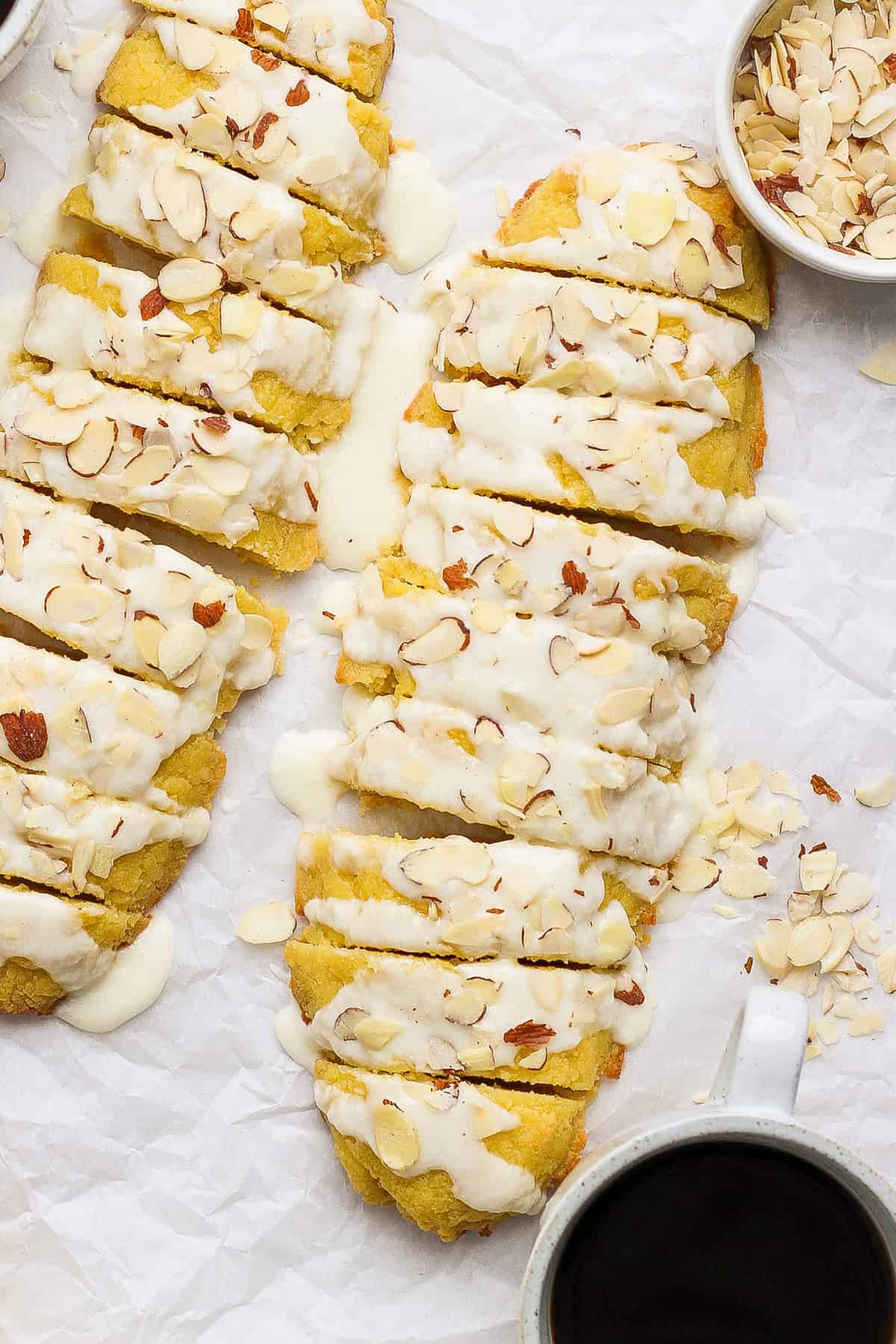 Slices of bread with white icing and almond flakes on parchment paper, alongside a bowl of almonds and a cup of black coffee.