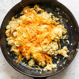 Scrambled eggs with shredded cheese in a black skillet on a light-colored surface.