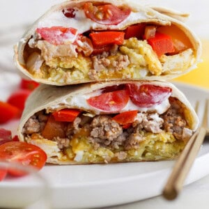 Two halves of a breakfast burrito filled with eggs, sausage, tomatoes, and topped with sour cream are stacked on a white plate.