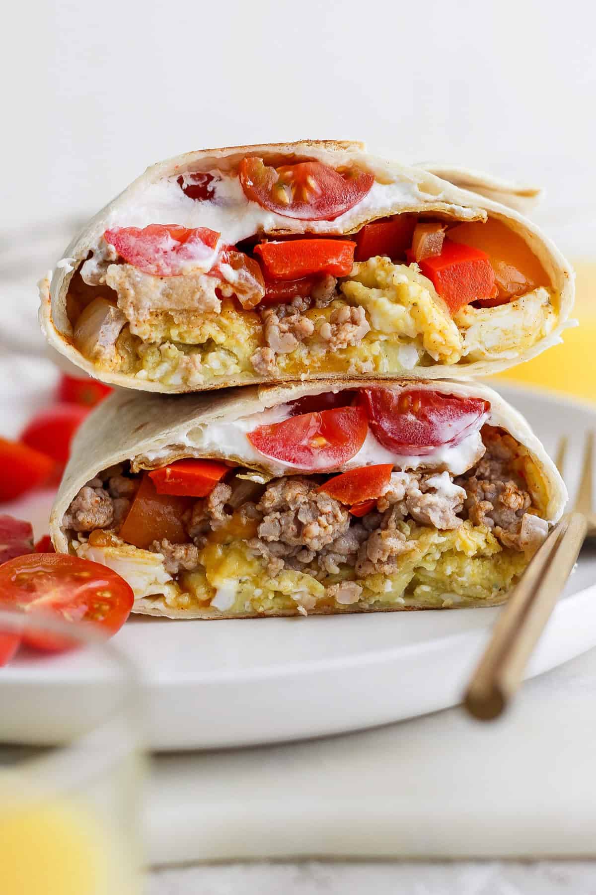 Two halves of a breakfast burrito filled with eggs, sausage, tomatoes, and topped with sour cream are stacked on a white plate.