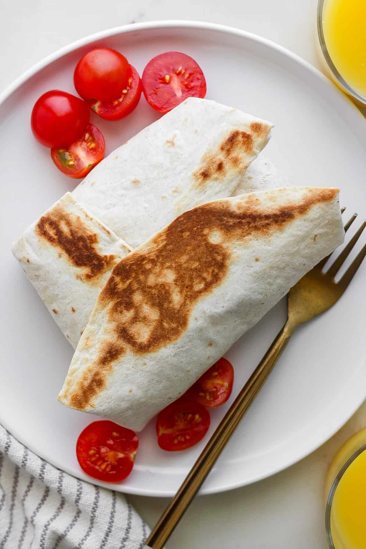 Two grilled burritos on a white plate surrounded by sliced cherry tomatoes, with a fork placed beside them.