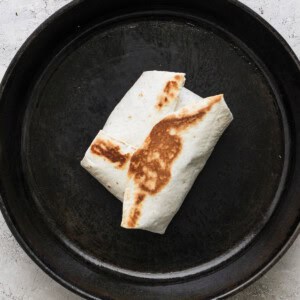 A folded and grilled burrito in a black cast iron pan on a gray surface.