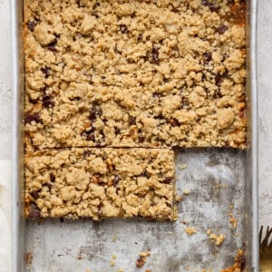 A metal baking pan filled with crumbly dessert bars. Two bars have been cut out, revealing a layered filling, while the rest of the dessert remains uncut. A fork and a napkin are nearby.