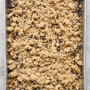 A rectangular baking tray filled with an unbaked dessert mixture featuring a crumbly oat and nut topping, placed on a textured light-colored surface.