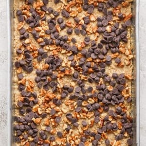 A silver baking pan containing an uncut dessert topped with chocolate chips and chopped nuts.